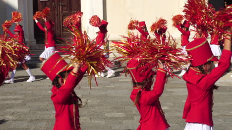 Chicas-Jóvenes-En-Una-Banda-De-Majorette,-Caminando-Con-Uniformes-Rojos-Y-Saludando-Con-Pompones.