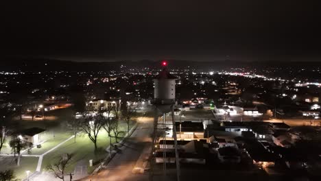 Drone-Volando-En-órbita-Alrededor-De-La-Torre-Del-Tanque-De-Agua-De-Una-Pequeña-Ciudad-Por-La-Noche-Junto-Al-Parque-Verde-De-La-Ciudad