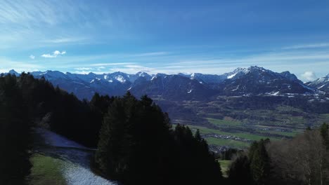 Drone-fly-next-to-forest-and-reveal-scenic-winter-mountain-landscape-with-snowcapped-mountains-in-background-and-small-town-in-Austria