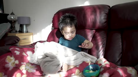 British-Asian-18-month-old-baby-boy-sits-on-a-sofa,-happily-eating-sliced-apples-with-morning-sunlight-hitting-sofa