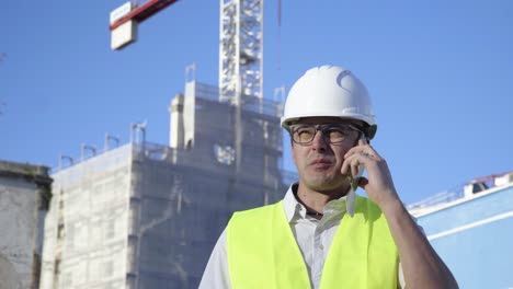 On-the-construction-site,-a-young-engineer-wearing-glasses-and-a-white-helmet-solves-problems-on-the-phone