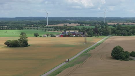 Traktor-Fährt-Auf-Der-Straße-Entlang-Ackerland-Mit-Windturbinen-Im-Hintergrund