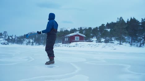 The-Man-is-Drilling-a-Hole-for-Ice-Fishing-Amidst-Fierce-Gusts-of-Wind-in-Bessaker,-Trondelag-County,-Norway---Static-Shot