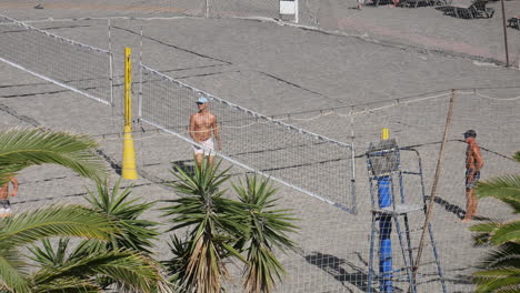 Male-players-playing-a-beach-volleyball-match,-surrounded-by-palm-trees-and-sand,-under-a-sunny-sky,-men-engaging-in-outdoor-sports