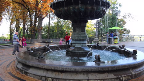 Schöner-Springbrunnen-In-Einem-Park-In-Der-Nähe-Der-Glasbrücke-Im-Stadtzentrum-Von-Kiew,-Ukraine,-Menschen-Genießen-Die-Natur-Mit-Großen-Bäumen,-4k-Aufnahme