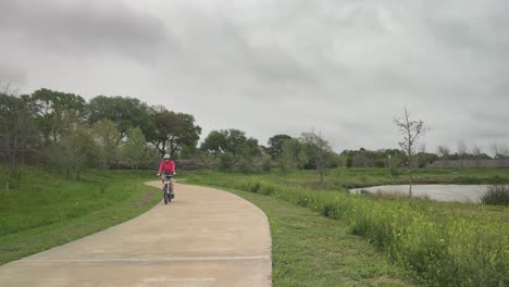 Una-Mujer-Mayor-Monta-Su-Bicicleta-En-Un-Día-Nublado-Y-Ventoso-De-Primavera-En-Exploration-Green-En-Clear-Lake,-Houston,-Texas