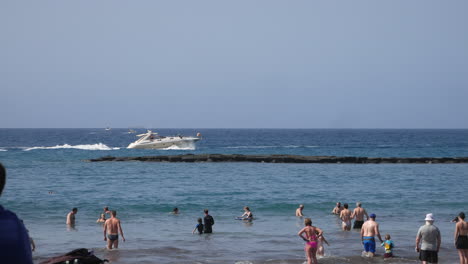 Urlauber-Schwimmen-Und-Waten-Im-Meer,-Während-Eine-Elegante-Weiße-Yacht-Vorbeirauscht-Und-Ein-Steinwellenbrecher-An-Einem-Sonnigen-Tag-Mit-Einem-Lebensstil-Des-Wohlstands-Sanfte-Wellen-Erzeugt