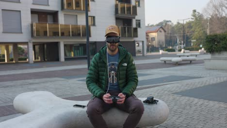 Drone-pilot-operator-sitting-on-city-bench-with-cap-controlling-DJI-Avata-quadcopter