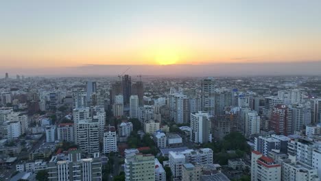 Drone-forward-over-skyscrapers-city-Santo-Domingo-with-orange-lit-sky,-sunset