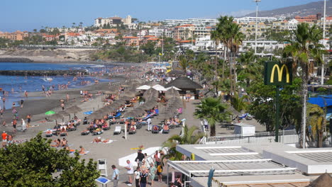 Der-Belebte-Strand-Von-Costa-Adeje-Auf-Teneriffa-Wimmelt-Von-Sonnenanbetern,-Strohgedeckten-Sonnenschirmen,-Sonnenbaden,-Blauem-Meerwasser-Und-Palmen-Vor-Der-Kulisse-Von-Resorthotels