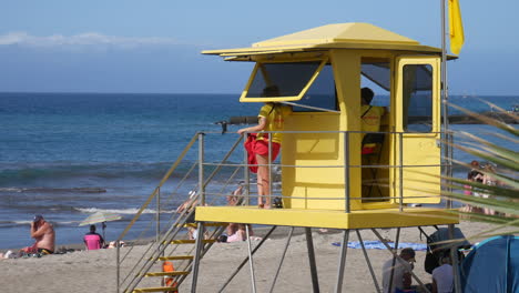 Lifeguards-in-yellow-tower-watch-over-sunbathers-and-swimmers-on-a-sunny,-palm-lined-sandy-beach-with-gentle-waves