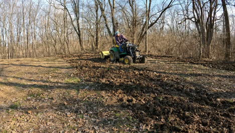 Hombre-Conduciendo-Un-Tractor-Utilitario-Remolcando-Un-Motocultor-Para-Aflojar-El-Suelo-En-Una-Parcela-De-Comida-Para-Ciervos-Entre-La-Madera-Y-El-Campo-A-Principios-De-La-Primavera