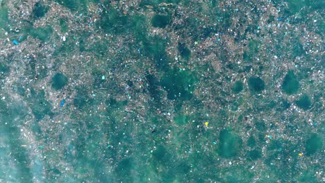 Top-down-quickly-ascending-drone-shot-of-polluted-water-filled-with-trash-floating-over-dead-coral-reef-in-the-turqouise-tropical-water-of-Bali-Indonesia