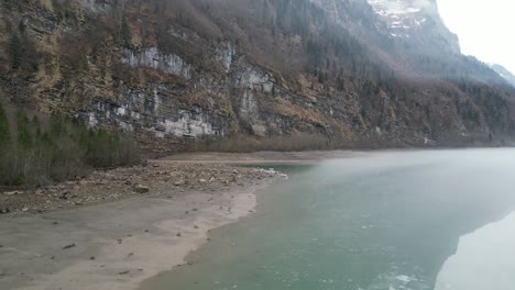 Dolly-along-sandy-grey-shoreline-with-boulders-on-edge-of-forest-canopy,-water-reflects-imposing-large-sheer-cliffs-and-mist