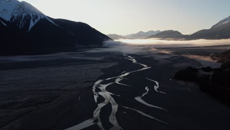 Aerial-view-of-the-braided-Bealey-river-in-shadow-in-Arthurs-pass-at-sunrise-in-a-valley-surrounded-by-mountains,-flying-towards-the-mountains-and-the-sun
