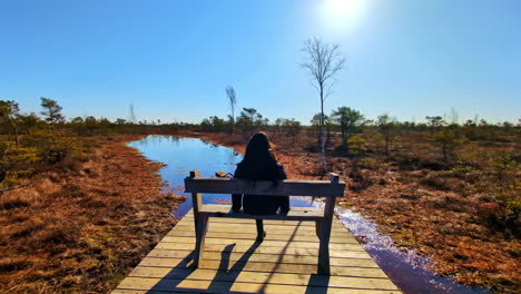 Solo-woman-relaxes-sitting-on-wooden-bench-standing-up-to-look-out-over-wetland-plains-at-midday