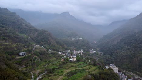 Misty-mountain-village-nestled-in-lush-valley,-overcast-day,-aerial-view