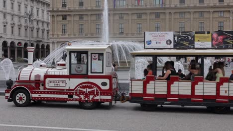 Red-and-white-tourist-train-moving-through-Piazza-De-Ferrari-in-Genoa,-Italy-with-traditional-buildings-and-tall-water-fountains-in-the-background