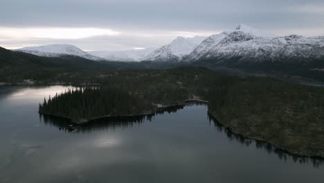 A-serene-half-frozen-lake-in-norway-with-snowy-mountains-at-dusk,-aerial-view