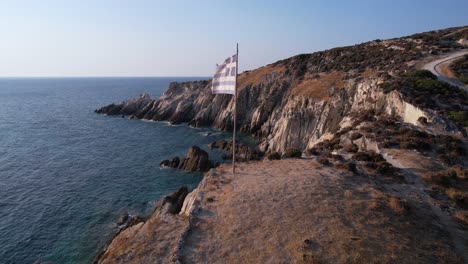 Vista-Aérea-De-La-Bandera-Nacional-Griega-Ondeando-En-El-Poste-Y-El-Acantilado-Sobre-El-Mar-Egeo.