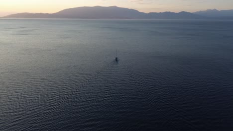 Single-boat-floating-on-ocean-with-riplles,-tilted-aerial-shot-towards-sun-lit-horizon-and-mountainous-coastline