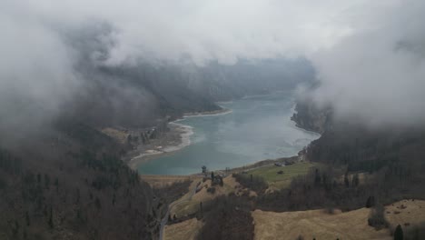 Drone-descends-between-fluffy-grey-white-clouds-to-reveal-frozen-lake-and-dam-in-European-mountains