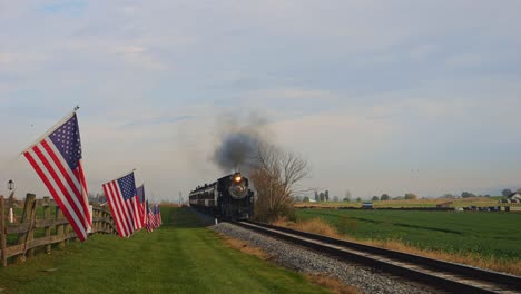 Una-Vista-De-Una-Sola-Vía-Férrea,-Con-Un-Tren-De-Pasajeros-A-Vapor-Pasando,-Con-Una-Valla-Con-La-Bandera-De-Estados-Unidos,-Ondeando-Suavemente-En-El-Viento-En-Un-Soleado-Día-De-Otoño