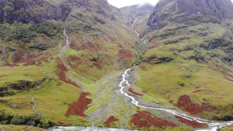 Luftaufnahme-Der-Dramatischen-Glencoe-Tallandschaft-In-Schottland-Mit-Fluss,-Der-Den-Berg-Hinunterfließt