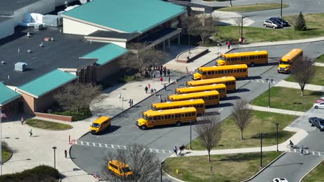 Toma-Aérea-Con-Zoom-Que-Muestra-Un-Grupo-De-Autobuses-Escolares-Amarillos-Esperando-En-El-área-De-Estacionamiento-Frente-A-La-Escuela-Americana