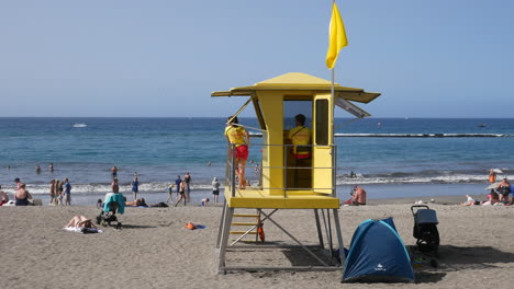 Cabaña-De-Salvavidas-Amarilla,-Monitores-Vigilantes,-Bañistas-Bañados-Por-El-Sol,-Olas-Suaves-Y-Serenidad-Costera-Bajo-Un-Cielo-Azul-Claro,-Resumen-Un-Día-De-Playa-Seguro