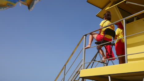 Lifeguards-in-red-and-yellow-attire-survey-the-beach-from-atop-a-yellow-tower-against-a-clear,-deep-blue-sky,-vigilant-over-seaside-safety