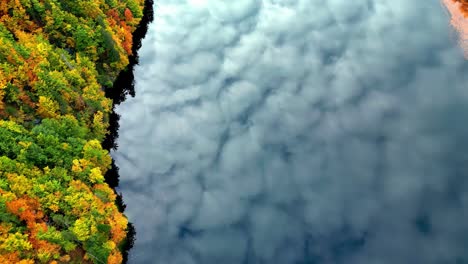 Reflction-of-clouds-in-autumn-mountain-lake,-winding-water-snaking-through-a-dense-forest,-with-a-blue-sky-and-puffy-white-clouds-above-lake-in-Alps