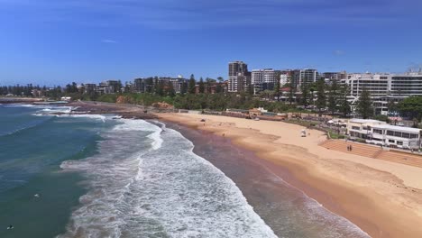 Antena-Sobre-Surfistas-Y-Hacia-Banderas-Que-Salvan-Vidas-En-La-Playa-De-North-Wollongong-Con-Un-Rascacielos-Más-Allá