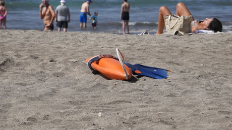 Orange-lifebuoy-and-blue-fins-lie-on-the-sandy-beach,-symbols-of-aquatic-safety-and-adventure-among-relaxed-beachgoers-under-the-summer-sun-with-a-man-walking-in-sandals