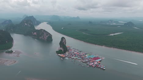 Drone-shot-captures-the-small-floating-village-of-Kon-Panyi-nestled-amidst-lush-greenery,-surrounded-by-towering-mountains-and-a-sparkling-blue-sea