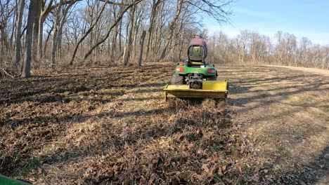 POV-following-a-man-on-utility-tractor-towing-a-rototiller-to-loosen-soil-in-a-deer-food-plot-between-timber-and-field-in-early-spring