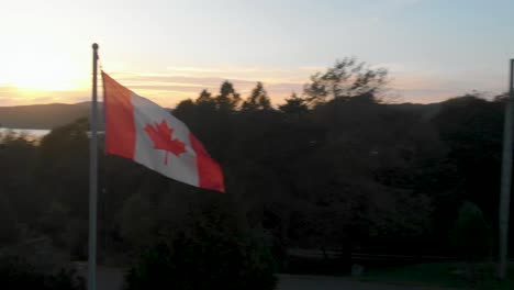 Halbumlaufende-Luftaufnahme-Einer-Kanadischen-Flagge-Auf-Einem-Im-Wind-Wehenden-Mast-Mit-Einem-Atemberaubenden-Sonnenuntergang-Hinter-Der-Baumgrenze