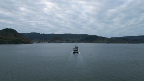A-ferry-crossing-calm-waters-near-nesvik-with-hjelmeland-backdrop,-cloudy-skies,-aerial-view