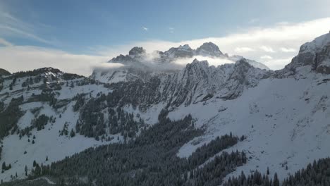 Antena-Panorámica-Que-Establece-Una-Plataforma-Rodante-De-Crestas-Rocosas-Irregulares-Y-Un-Bosque-De-Pinos-Cubierto-De-Nieve-Con-Nubes-Que-Se-Reúnen-Alrededor-De-La-Montaña