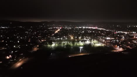 Drone-Pan-Horizontal-Volando-Por-Una-Pequeña-Ciudad-Por-La-Noche-Con-Calles-Verdes-Del-Parque-De-La-Ciudad-Con-Autos