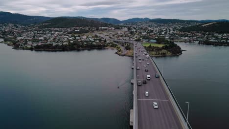 Toma-Aérea-De-Un-Puente-Con-Vehículos-Circulando-Sobre-él-En-Un-Día-Nublado-Con-Paisaje-Urbano-Al-Fondo