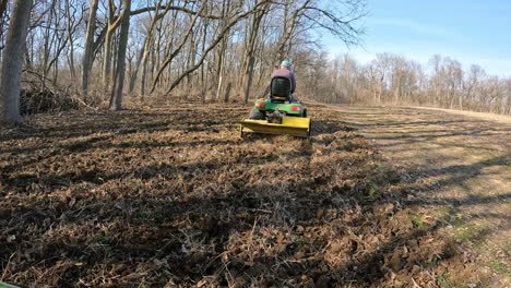 POV-following-a-man-on-utility-tractor-towing-a-rototiller-to-loosen-soil-in-a-wildlife-food-plot-between-wood-and-field-in-early-spring