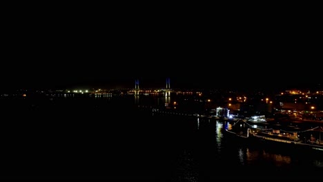 Night-view-of-a-bridge-illuminated-in-blue-with-city-lights-and-moored-boats,-reflected-on-water