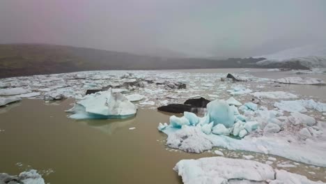 Low-flyover-of-icebergs-floating-in-muddy-glacier-lagoon-on-foggy-day-in-Iceland