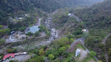 A-lush-green-valley-with-a-winding-river,-roads-and-buildings,-daylight,-aerial-view