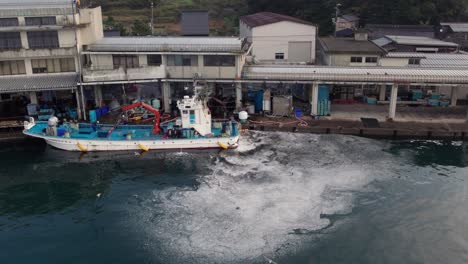 fishing-boat-sails-at-Japanese-Kyotango-dock-seagulls-birds-fly-by-aerial-drone-blue-Japan-Kansai-ship-industry-landscape,-Kyoto-countryside