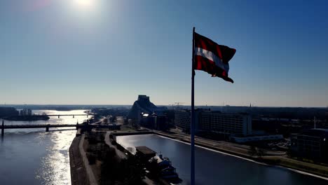 La-Bandera-Letona-En-La-Presa-De-Ganību,-Ondeando-Sobre-El-Río-Daugava,-Deleita-A-Los-Residentes-Del-Centro-De-La-Ciudad-De-Riga.