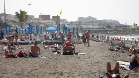 überfüllte-Sandstrandszene-Mit-Gelber-Flagge,-Sonnenanbetern,-Blauen-Sonnenschirmen,-Sanfter-Brandung-Und-Urlaubsstimmung-Unter-Einem-Dunstigen-Blauen-Himmel-An-Der-Costa-Adeje-Auf-Teneriffa