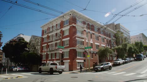 Esquina-De-La-Calle-Del-Centro-De-San-Francisco-Mientras-Los-Automóviles-Pasan-Por-Viajeros-Que-Caminan-Debajo-De-Las-Líneas-De-Cable-Ferroviario