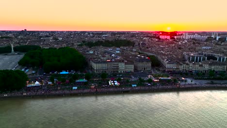 Gente-Reunida-En-La-Orilla-Del-Río-Garona-Con-Noria-En-Place-Des-Quinconces-Burdeos-Francia-Durante-La-Puesta-De-Sol,-Panorámica-Aérea-Izquierda-Revela-Toma